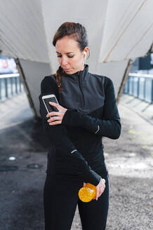 Weibliche Sportlerin benutzt ihr Smartphone über ein Armband, während sie auf dem Gehweg steht - JMPF00418