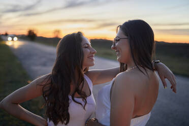 Smiling female friends looking at each other while standing on road against sky at sunset - ZEDF03786