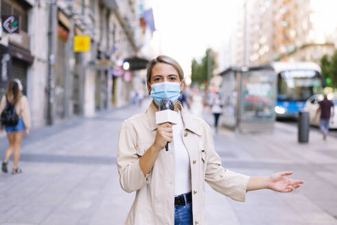Female reporter wearing mask talking over microphone on street in city - JCMF01476