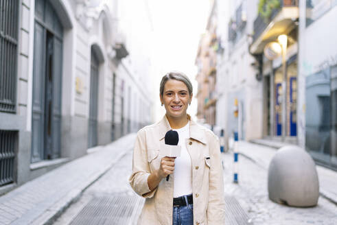 Eine Journalistin spricht über ein Mikrofon, während sie auf einer Straße in der Stadt steht - JCMF01487