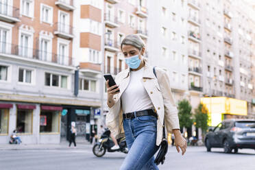 Junge Frau mit Maske, die ein Mobiltelefon benutzt, während sie auf einer Straße in der Stadt geht - JCMF01489