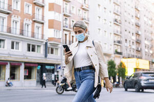 Young woman wearing mask using mobile phone while walking on street in city - JCMF01489