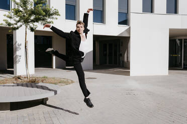 Carefree trans young man wearing suit dancing on footpath against building in city - TCEF01082