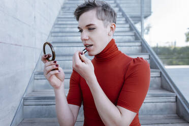 Close-up of trans young man applying lipstick while sitting on steps - TCEF01083
