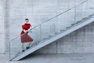 Trans young man wearing red dress standing on steps by railing - TCEF01085