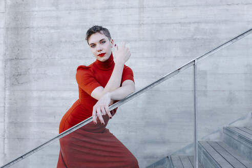 Confident trans man wearing red dress standing on steps against wall - TCEF01086