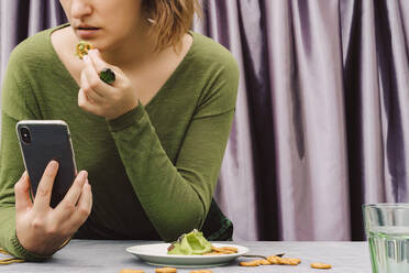 Nahaufnahme einer Frau, die eine Brezel mit Guacamole isst und dabei ihr Smartphone auf einem Tisch zu Hause benutzt - ERRF04434