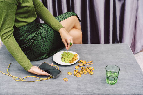 Frau hält Smartphone und isst Guacamole und Brezel, während sie zu Hause am Tisch sitzt - ERRF04439