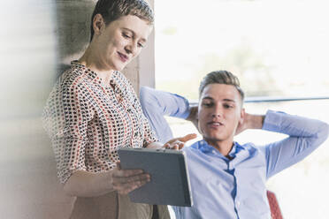 Eine Geschäftsfrau zeigt einem männlichen Mitarbeiter ein digitales Tablet, während sie mit ihm im Büro diskutiert - UUF21500