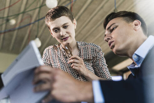 Männlicher Berufstätiger, der mit seiner Partnerin im Büro über ein digitales Tablet diskutiert - UUF21515