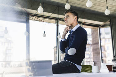 Thoughtful businessman with hand on chin looking away while sitting over desk in office - UUF21517