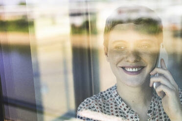 Close-up of smiling businesswoman talking over smart phone in office seen through window - UUF21521