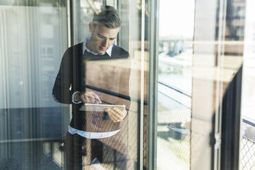 Male professional working over digital tablet while standing in office seen through window - UUF21524