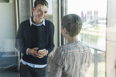 Smiling businessman holding smart phone talking with female coworker in office seen through window - UUF21527