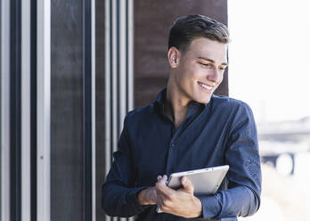 Lächelnder männlicher Fachmann, der ein digitales Tablet hält, während er am Geländer eines Balkons steht - UUF21536