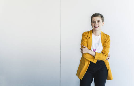 Lächelnde weibliche Fachkraft in gelbem Blazer, die wegschaut, während sie im Büro an der Wand steht - UUF21554