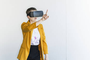 Geschäftsfrau mit Blazer, die durch einen Virtual-Reality-Simulator schaut, während sie im Büro an der Wand steht - UUF21558