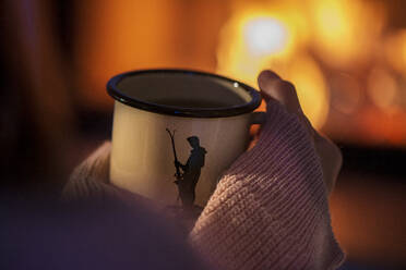 Close-up of young woman hands holding tea cup in front of fireplace at home - LBF03234