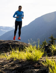 Vorderansicht einer Sportlerin in Sportkleidung, die an einem sonnigen Abend auf dem Lande auf Felsen in der Nähe von Bergen läuft - CAVF89287
