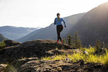Ganzkörper-Sportlerin in Sportkleidung läuft auf Felsen in der Nähe von Bergen an einem sonnigen Abend auf dem Lande - CAVF89288