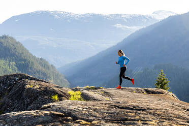 Side view of female athlete in sportswear trail running on rocks backed by snowy mountains on sunny evening in countryside - CAVF89289