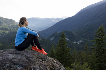 Kräftige Sportlerin, die beim Fitnesstraining auf dem Lande vor einem bewaldeten Bergrücken sitzt und eine Pause vom Trailrunning einlegt - CAVF89293