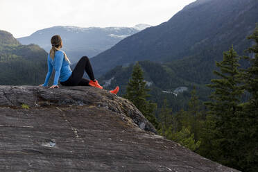 Weibliche Athletin sitzt und macht eine Pause vom Trailrunning gegen einen bewaldeten Bergrücken während eines Fitnesstrainings auf dem Lande - CAVF89294