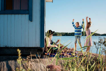 Woman sat on a rock playing guitar whilst her kids dance in the sun - CAVF89297