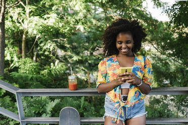 Lächelnde Frau, die mit ihrem Smartphone SMS schreibt, steht an einem Geländer im Park - XLGF00576