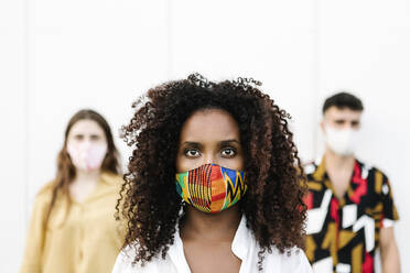 Young woman wearing face mask with friends standing against wall - XLGF00591