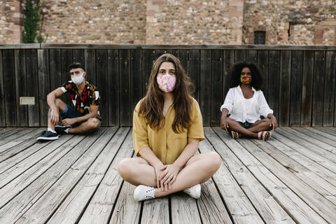 Friends wearing protective face mask while sitting on wooden floor during covid-19 stock photo