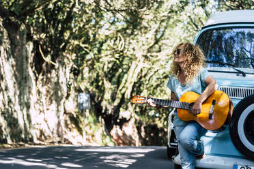 Mature woman wearing sunglasses playing guitar while standing by motor home on road - SIPF02195