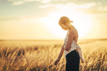 Junge Frau mit schulterfreiem Oberteil steht in einem Feld gegen den Himmel bei Sonnenuntergang - ACPF00807