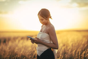 Junge Frau mit schulterfreiem Oberteil, die ein Smartphone benutzt, während sie in einer Landschaft bei Sonnenuntergang steht - ACPF00809