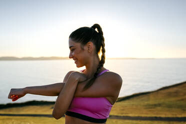 Smiling young woman stretching hands while standing against sea at sunset - MGOF04465