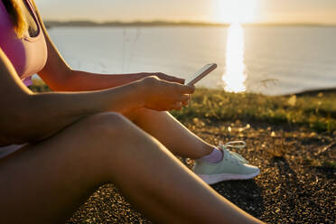 Nahaufnahme einer jungen Frau, die ihr Smartphone benutzt, während sie bei Sonnenuntergang auf einer Straße am Meer sitzt - MGOF04469