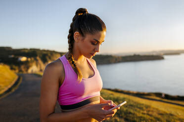 Junge Frau, die ihr Smartphone benutzt, während sie auf einer Klippe gegen den klaren Himmel bei Sonnenuntergang steht - MGOF04473