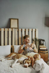 Ein junger blonder Junge mit einer Tasse Kaffee und seinem Hund im Bett - CAVF89422
