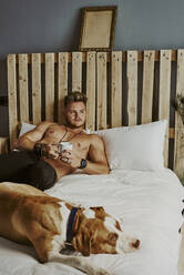 Ein junger blonder Junge mit einer Tasse Kaffee und seinem Hund im Bett - CAVF89424