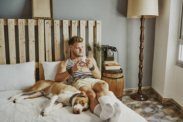 Ein junger blonder Junge mit einer Tasse Kaffee und seinem Hund im Bett - CAVF89425