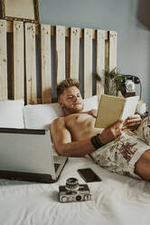 A man reads a book while working with his cell phone and laptop in a hotel bed - CAVF89428