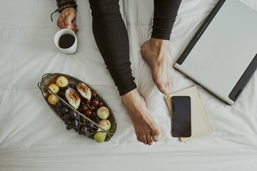 Feet and legs of a man in bed next to a computer, fruit, coffee, - CAVF89439