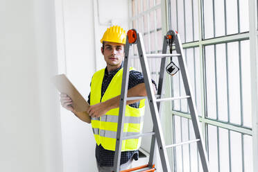 Architekt mit Arbeitshelm und Jacke steht auf einer Leiter in einem im Bau befindlichen Büro - GIOF08844