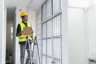 Man wearing work helmet and jacket standing on ladder by window at construction site - GIOF08846