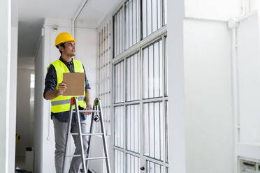 Mann mit Arbeitshelm und Jacke steht auf einer Leiter am Fenster auf einer Baustelle - GIOF08846