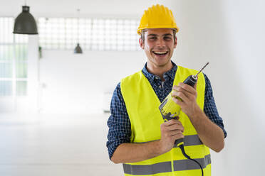 Smiling man holding drill machine while standing at office under construction - GIOF08847