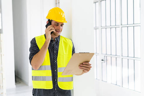 Lächelnder Architekt, der einen Notizblock hält, während er in einem im Bau befindlichen Büro mit einem Mobiltelefon spricht - GIOF08849