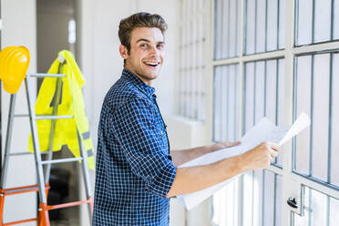 Smiling man holding blueprint while standing by window at construction site - GIOF08858