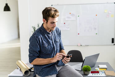 Young man using mobile phone while sitting on desk at office - GIOF08878