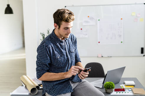 Junger Mann, der ein Mobiltelefon benutzt, während er auf dem Schreibtisch im Büro sitzt - GIOF08878